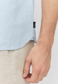 Lichtblauwe kort--mouw shirt van zachte stof, met een zijslit en een zwart merketiket aan de zoom, gecombineerd met beige broek.