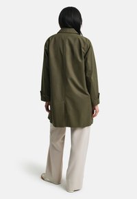 Žena s dlouhými tmavými vlasy, oblečená v olivově zeleném dlouhém kabátu, béžových kalhotách s širokými nohavicemi a světlých botách, stojí s obratem zády.