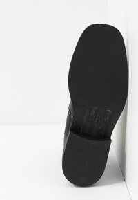 Zapato de cuero negro con una textura suave y punta redondeada; presenta una suela negra mate y sutiles remaches decorativos a lo largo del borde superior.
