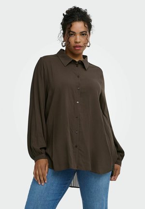 Donna con capelli ricci indossa una camicia oversize marrone scuro con bottoni e jeans azzurri, in piedi davanti a uno sfondo chiaro e semplice.