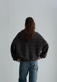 Dikke zwarte trui met kabelbreipatroon, oversized pasvorm en ribgebreide manchetten, gecombineerd met lichtblauwe jeans. De textuur lijkt zacht en knus.