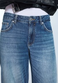 Modré džínové kalhoty s vybledlým efektem a rovnými nohavicemi. Mají pět kapes, viditelné švy a kovové zapínání na knoflík.
