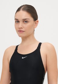 Črn kopalke z okroglim izrezom in tankimi naramnicami. Imajo bel Nike logotip na prsih in diskretne šive ob straneh.