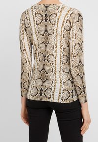 Długi rękaw w bluzce z brązowo-kremowym wzorem węża, z okrągłym dekoltem i prążkowaną fakturą, noszony z czarnymi spodniami.