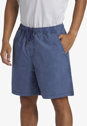Homme portant un short bleu à cordon et un t-shirt blanc, avec une main dans la poche et des détails de couture visibles sur le short.