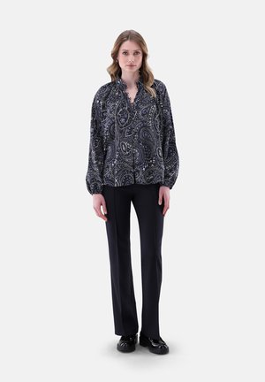 Donna in piedi che indossa una blusa paisley bianca e nera, pantaloni neri a gamba dritta e mocassini neri su uno sfondo bianco.