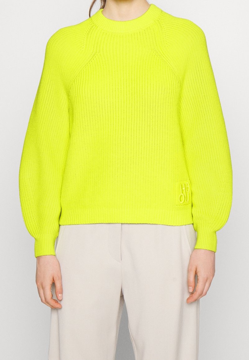 Personne portant un pull en maille jaune néon vif avec des manches ballon et un pantalon beige clair, sur un fond uni.