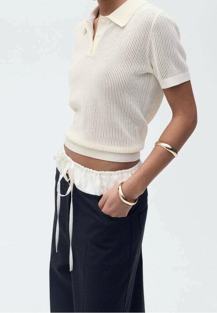 Femme portant un polo en maille blanc à manches courtes, un pantalon noir taille haute avec un cordon blanc à la taille, et des bracelets dorés au poignet.