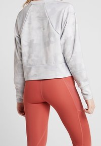 Felpa cropped grigia chiara con un sottile motivo camouflage, dotata di maniche raglan e una consistenza morbida, abbinata a leggings color ruggine.