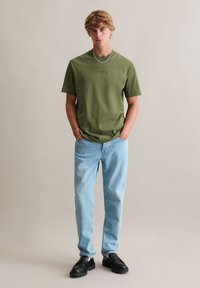 Oliwkowa zielona bawełniana koszulka z okrągłym dekoltem, zestawiona z jasnoniebieskimi dżinsami i czarnymi masywnymi loafersami. Prosty, luźny krój i lekka faktura.