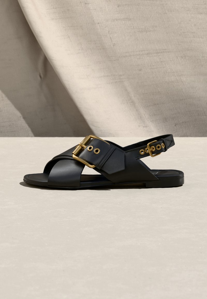 Μαύρο δερμάτινο σανδάλι με φαρδιές σταυρωτές λουρίδες, χρυσά πόρπες και λουρί που πιάνει πίσω, σε ουδέτερη επιφάνεια με φόντο  από ύφασμα.