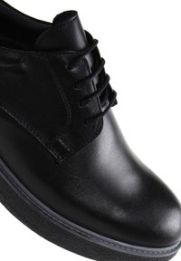 Zapato de piel negra con un acabado suave, punta redondeada y suela texturizada en contraste. Presenta cordones clásicos y detalles de costura.