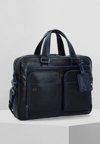 Borsa in pelle nera con cuciture blu, due tasche anteriori, doppi manici e un'etichetta rimovibile. Dotata di una tracolla staccabile.