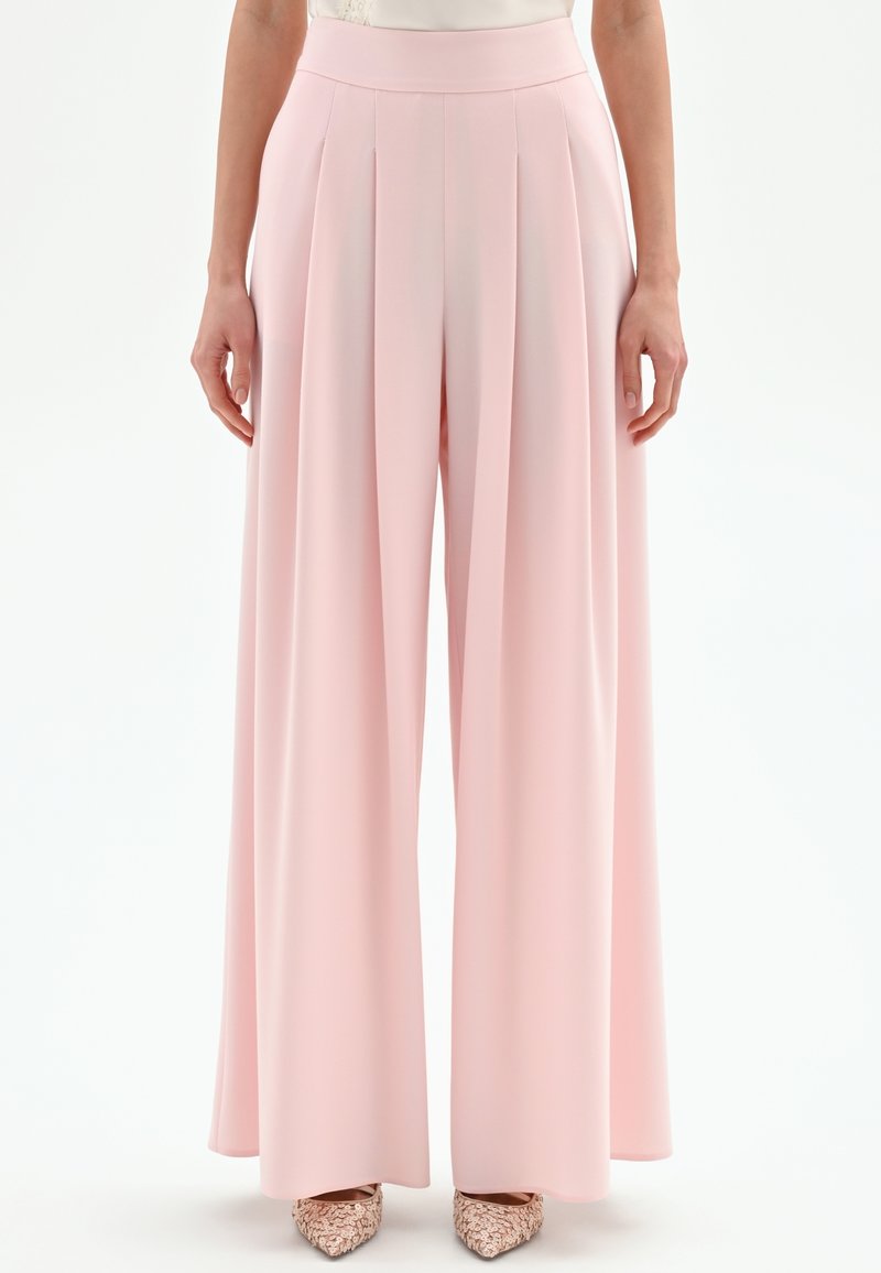 Pantalones anchos de pierna ancha en rosa claro con pliegues delanteros, cintura alta y textura de tela suave. Presentan un ajuste holgado y un diseño de largo completo.