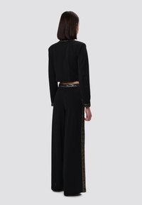 Zwarte cropped jas met lange mouwen, gecombineerd met brede zwarte broeken met een gouden geometrisch patroon langs de zijkant.