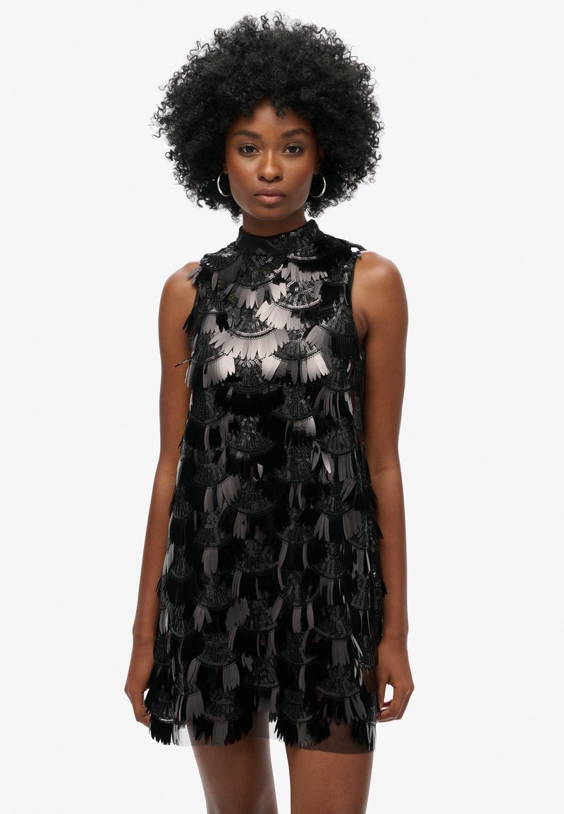 Robe noire sans manches avec des détails en franges superposées, dotée d'un col haut et d'une texture lisse. Le tissu a une finition brillante.