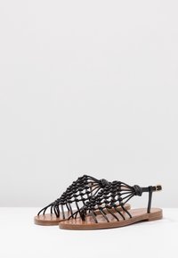Sandales noires tissées avec une semelle en bois plate, présentant des détails en corde complexes et une bride à la cheville avec boucle. Texture lisse et design minimaliste.