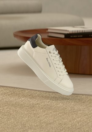 Weißer Marc O'Polo Sneaker mit schwarzem Fersen-Detail und Schnürsenkeln, platziert neben einem runden Holztisch mit aufgestapelten Büchern in einem in neutralen Tönen gehaltenen Raum.