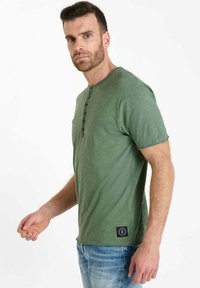 Green short-sleeve t-shirt in a textured material, featuring a round neckline and a button placket on the front. Black logo patch on the side.