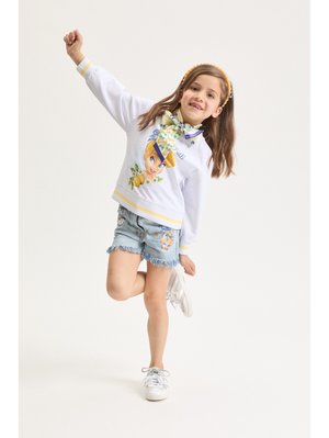 Ragazza che sorride e si equilibra su una gamba, indossando una felpa bianca con un volto stilizzato, pantaloncini di jeans e scarpe da ginnastica bianche, su uno sfondo neutro.