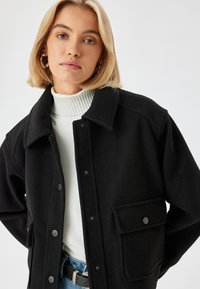 Schwarze Wollmischung Jacke mit Kragen, Druckknöpfen und einer Vordertasche. Getragen über einem weißen gerippten Rollkragenpullover, der einen lässigen Schnitt zeigt.