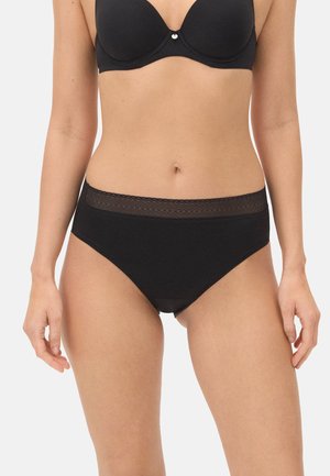 Mujer con sujetador negro y ropa interior negra de tiro alto con cintura de malla, de pie contra un fondo blanco.