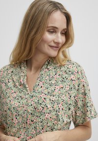 Blouse à motifs floraux avec un fond vert, des fleurs roses et oranges, et un décolleté en v avec des manches courtes. Texture en coton.