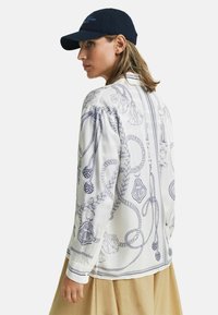 Weiße Bluse mit marineblauem Seilmotiv und dekorativen Elementen, mit langen Ärmeln, gerafften Schultern und geradem Saum. Kombiniert mit einer dunklen Mütze.