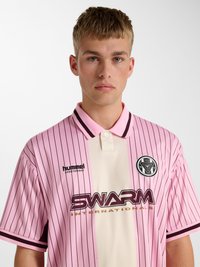 Młody mężczyzna z krótkimi blond włosami nosi różowo-kremową pasiastą koszulkę polo z logo "SWARM" oraz okrągłym emblematem na piersi.