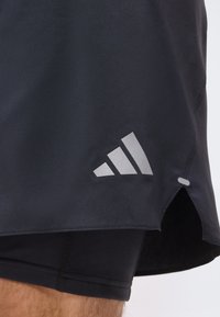 Shorts de sport noirs avec logo Adidas réfléchissant sur l'ourlet et une petite fente latérale, portés par-dessus des shorts de compression sur la jambe d'une personne.