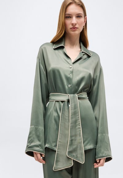 Γυναίκα που φοράει χαλαρό πουκάμισο από σατέν σε ελιά πράσινο χρώμα με φαρδιές μανσέτες και παντελόνι με ζώνη σε αντίστοιχο χρώμα, με φόντο ένα απλό γκρι πίσω μέρος.