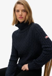 Joven con cabello rubio hasta los hombros, vistiendo un suéter de cuello alto de punto en azul oscuro, sentada en una silla de madera.