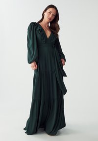 Dark green maxi dress made of soft fabric. Features long ruffled sleeves, a deep V-neck, and a tied waist with tiered bottom.