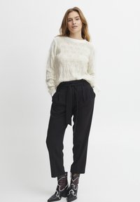 Cremefarbener Strickpullover mit strukturiertem Muster, kombiniert mit schwarzer High-Waist-Hose mit gebundenem Gürtel und schwarzen Ankle Boots mit weißen Akzenten.