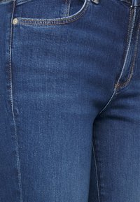 Jeans de mezclilla azul oscuro con una textura suave, diseño clásico de cinco bolsillos y costuras en contraste. Presentan un cierre de botón de metal y cremallera.