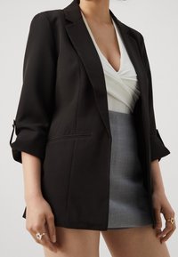 Femme portant un blazer noir, un haut blanc froncé et une mini-jupe grise, avec les manches du blazer retroussées et des bagues aux deux mains.