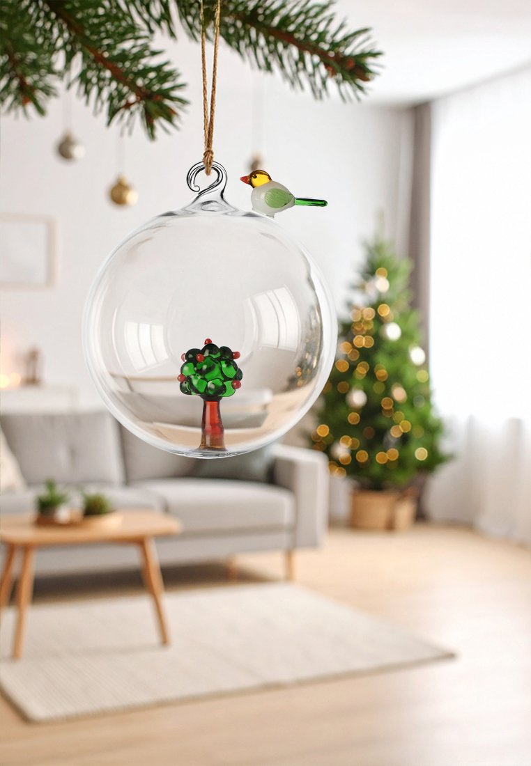Klarer Glasornament mit einem grünen Baum und roten Akzenten im Inneren, oben mit einem dekorativen Vogel versehen. Hängt an einem Jute-Band.