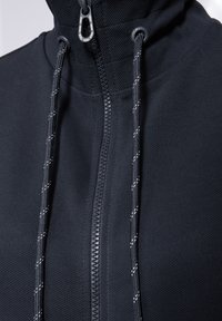 Felpa nera con zip, realizzata in tessuto testurizzato, con una zip centrale, cordini regolabili e dettagli riflettenti sui cordini.