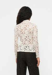 Top in pizzo a maniche lunghe di colore bianco sporco, caratterizzato da motivi floreali, un colletto alto e una forma aderente, abbinato a pantaloni neri.