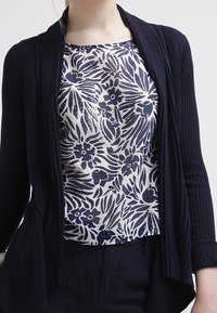 Mujer usando un cárdigan de punto de color azul marino sobre una camisa blanca con un patrón floral en azul marino, combinado con pantalones oscuros.