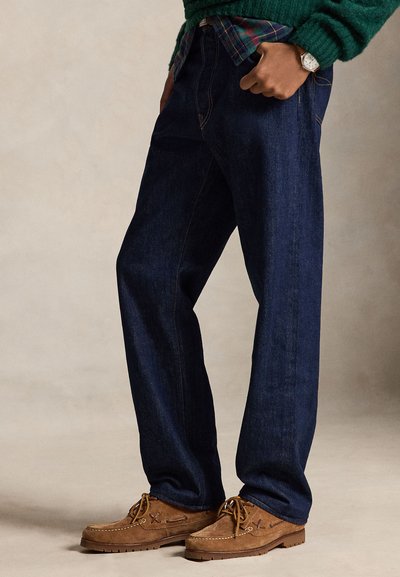 Persona con vaqueros azul oscuro, zapatos marrones de ante con cordones, suéter verde y camisa de cuadros con una mano en el bolsillo.
