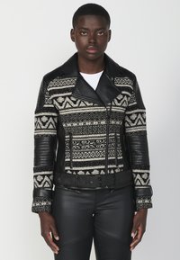 Giacca di pelle nera con tessuto a maglia decorato, caratterizzata da un ampio colletto, chiusure con zip e maniche testurizzate. Vita con cintura e dettagli decorativi.
