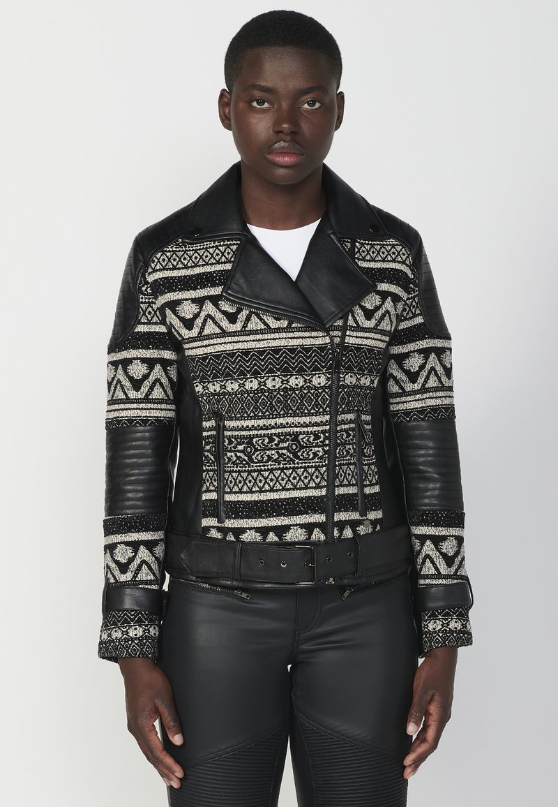 Giacca di pelle nera con tessuto a maglia decorato, caratterizzata da un ampio colletto, chiusure con zip e maniche testurizzate. Vita con cintura e dettagli decorativi.