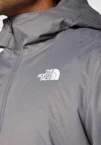 Grå vindjakke med tekstureret finish og lynlås krave. Har et hvidt logo fra The North Face på venstre brystområde.