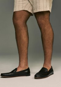 Sorte læder loafers med en struktureret overflade, der har en subtil detalje på toppen, båret med lysfarvede, strukturerede shorts.