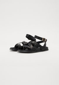 Black flat sandals made of smooth material, featuring multiple crossed straps and an ankle buckle. Simple design and minimalistic shape.