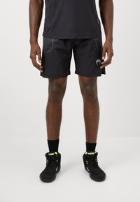 Mann med svarte treningsshorts og svarte høyhalse sneakers med neon grønn aksenter, stående mot en enkel hvit bakgrunn.