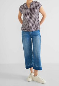 Blusa a maniche corte con scollo a V e motivo geometrico in rosso scuro e blu navy, abbinata a jeans in denim blu a gamba larga e sneakers bianche.