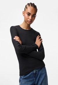 Must musträni varrukatega must ülikond, millel on ümmargune kaelus, koos kõrge talje ja siniste teksadega. Mudel poseerib käed ristis.