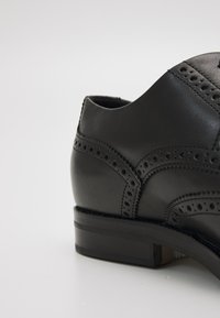 Juodas odinis batas su brogue detalėmis, lygiais ir raštuotais paviršiais, žemu kulnu ir apvaliu nosies dizainu.